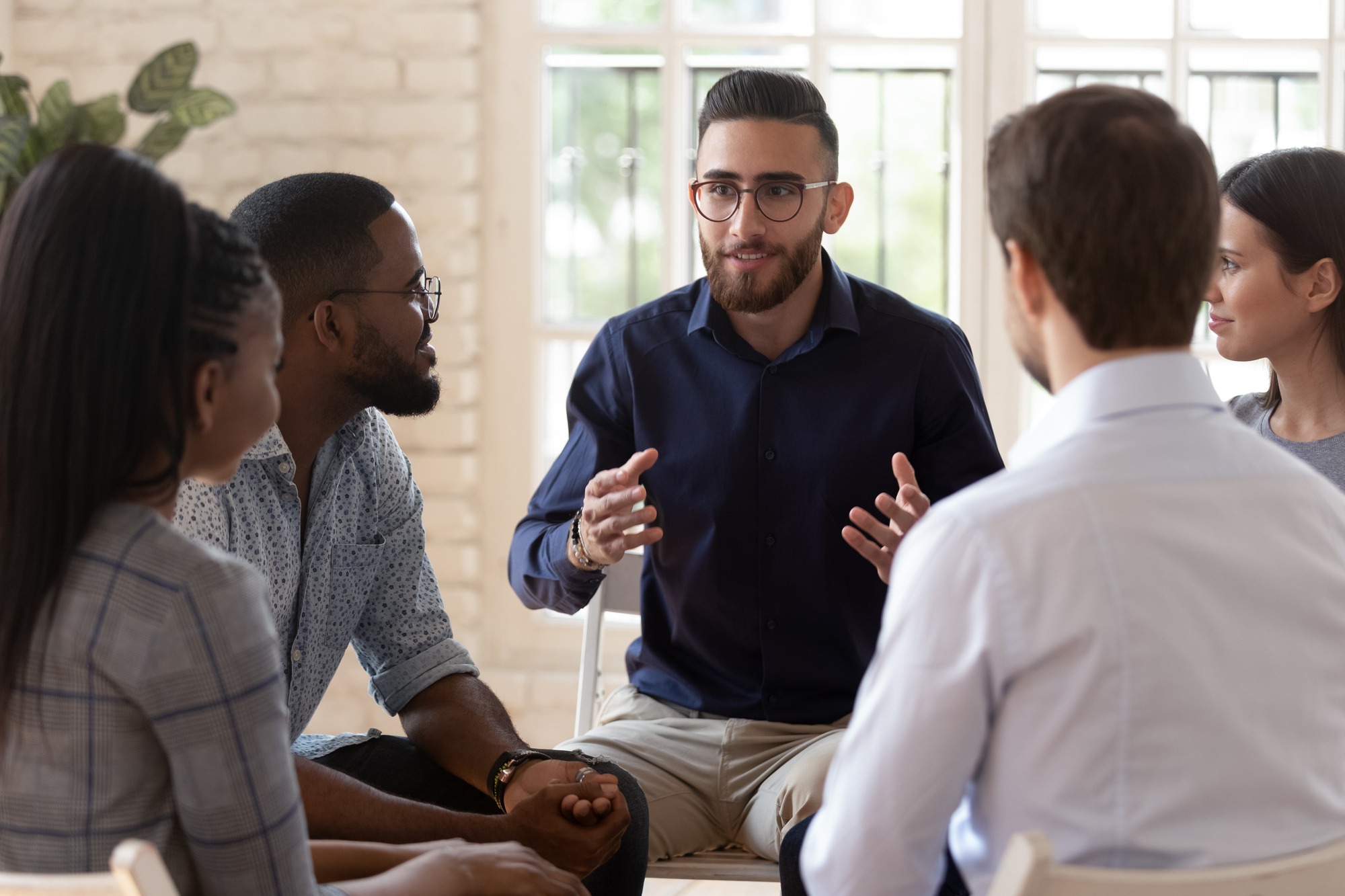 Happy Arabian millennial man speak at group motivational training share idea thought with friends, diverse young people sit in circle talk chat help cope with problems, psychological support concept.