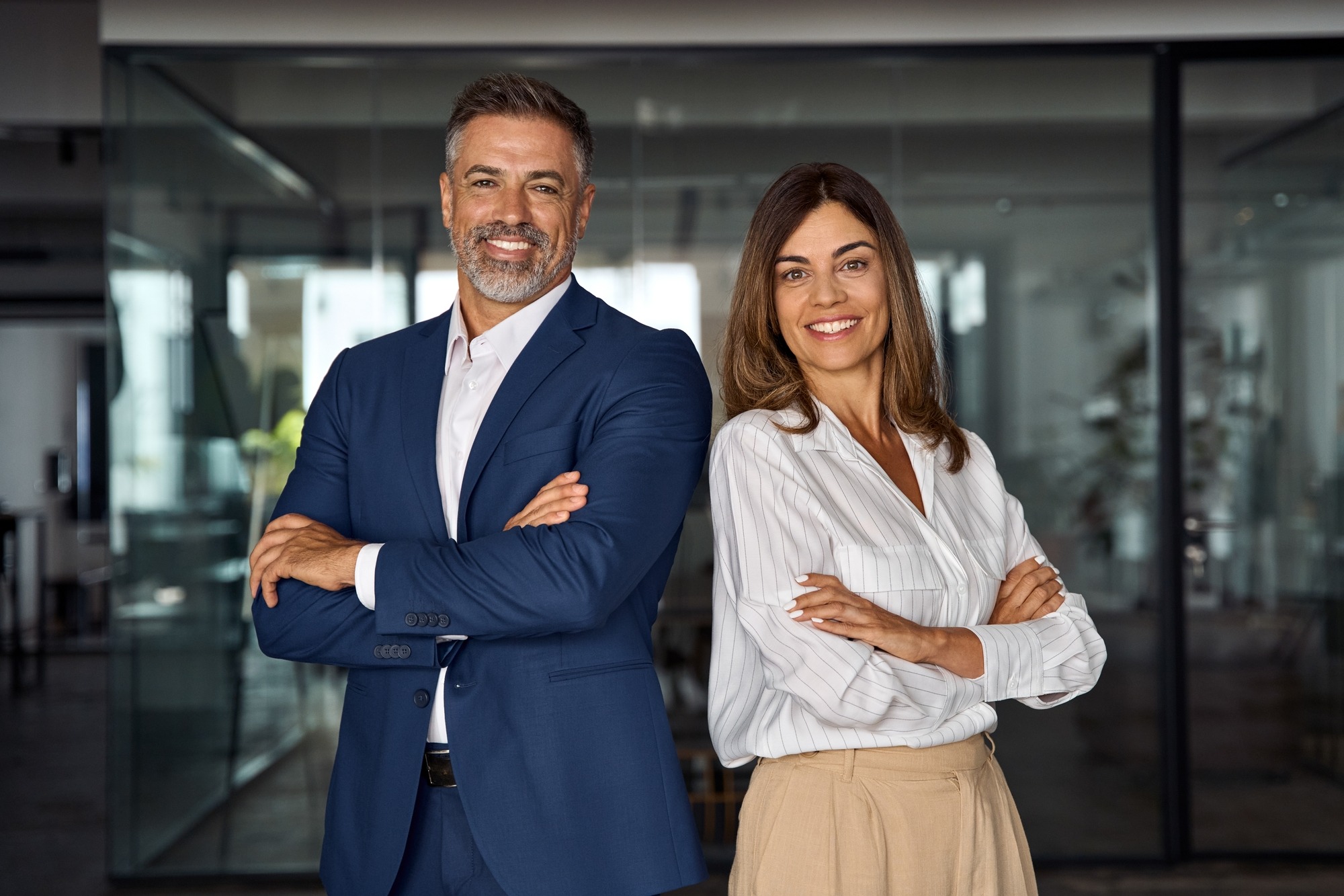 Portrait of smiling mature Latin or Indian business man and European business woman standing arms crossed in office. Two diverse colleagues, group team of confident professional business people.; Shutterstock ID 2343004303; purchase_order: Limited 5/13; job: ; client: ; other: